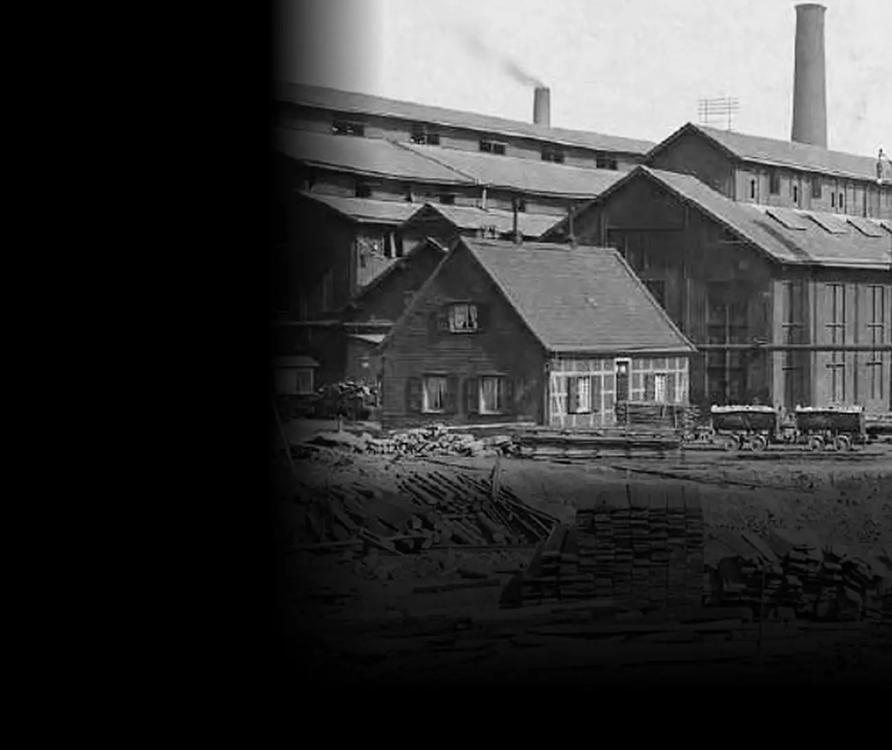 L'aciérie Krupp à Essen en 1908 : à gauche, une petite maison à colombages au toit de tuiles ; derrière et autour d'elle, de grandes halles, cinq hautes cheminées et une file de wagonnets ; au premier plan, un chantier jonché de planches.