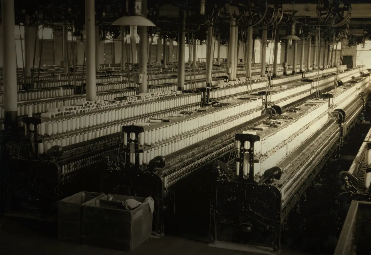 Une salle de filature mécanique de 1916 : des rangées de métiers à filer chargés de milliers de bobines blanches s'étendent jusqu'au fond, sous les poulies et les courroies du plafond. Personne n'est visible.