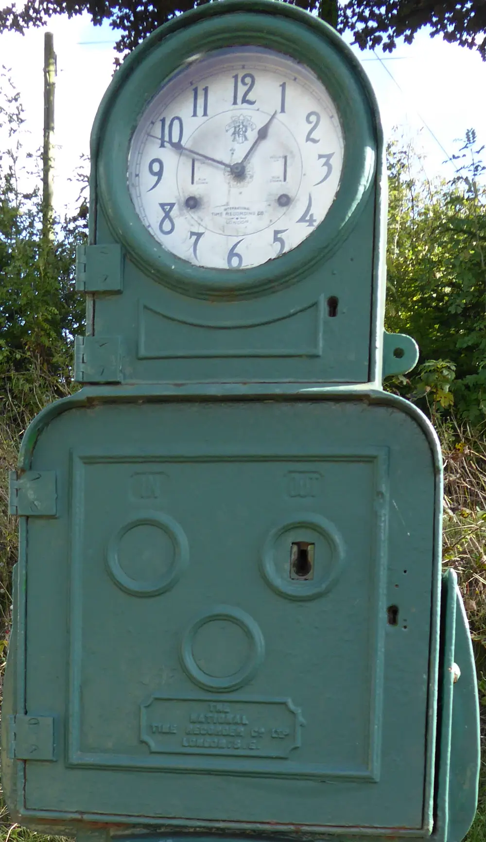 Une pointeuse ancienne en fonte peinte en vert : un cadran d'horloge en haut, et dessous deux ouvertures marquées IN et OUT, avec la plaque de la National Time Recorder Company de Londres.
