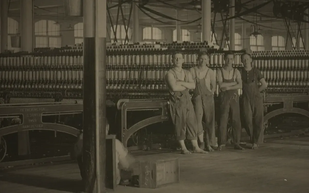 Photographie d'archive : la salle des métiers d'une filature en 1912, ses arbres de transmission au plafond, et quatre jeunes ouvriers pieds nus debout au milieu, bras croisés, qui regardent l'objectif.