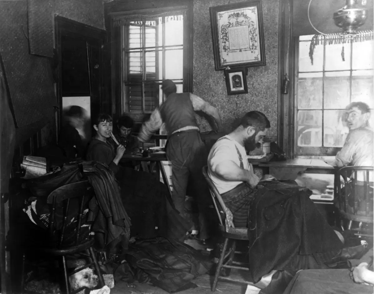 Une pièce d'habitation à New York : des hommes et des femmes cousent des vêtements assis sur des chaises de bois, des étoffes s'entassent sur le sol et sur les meubles, un chien regarde sous une chaise.