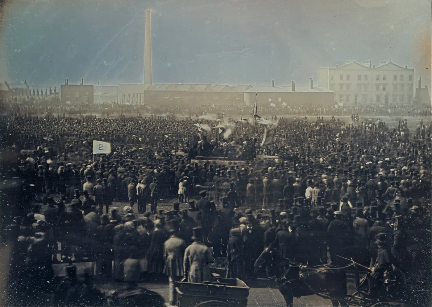 Une foule immense, en chapeaux, rassemblée sur un terrain découvert autour d'une estrade et de drapeaux ; au fond, des bâtiments bas et une haute cheminée d'usine.