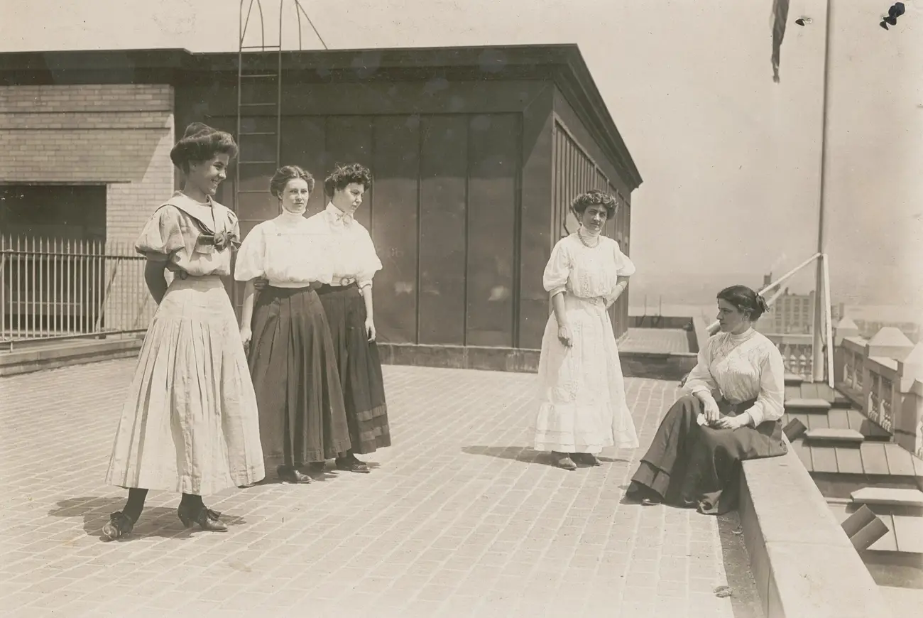 Photographie : cinq jeunes femmes en jupe longue et chemisier blanc, debout ou assises sur le toit-terrasse d'un immeuble de bureaux à New York.