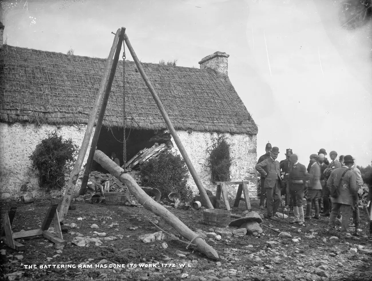 Photographie : une chaumière irlandaise dont le mur est éventré ; devant, un bélier de bois suspendu à un trépied ; à droite, un groupe d'hommes en chapeau melon et deux policiers casqués regardent.