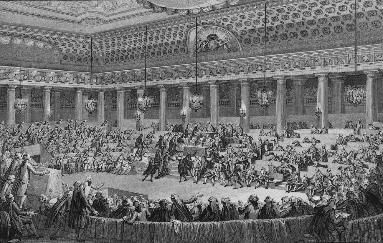 Gravure : la salle de l’Assemblée nationale à Versailles, la nuit du 4 août 1789 ; au centre, des députés se pressent vers le bureau du président pour renoncer à leurs privilèges.