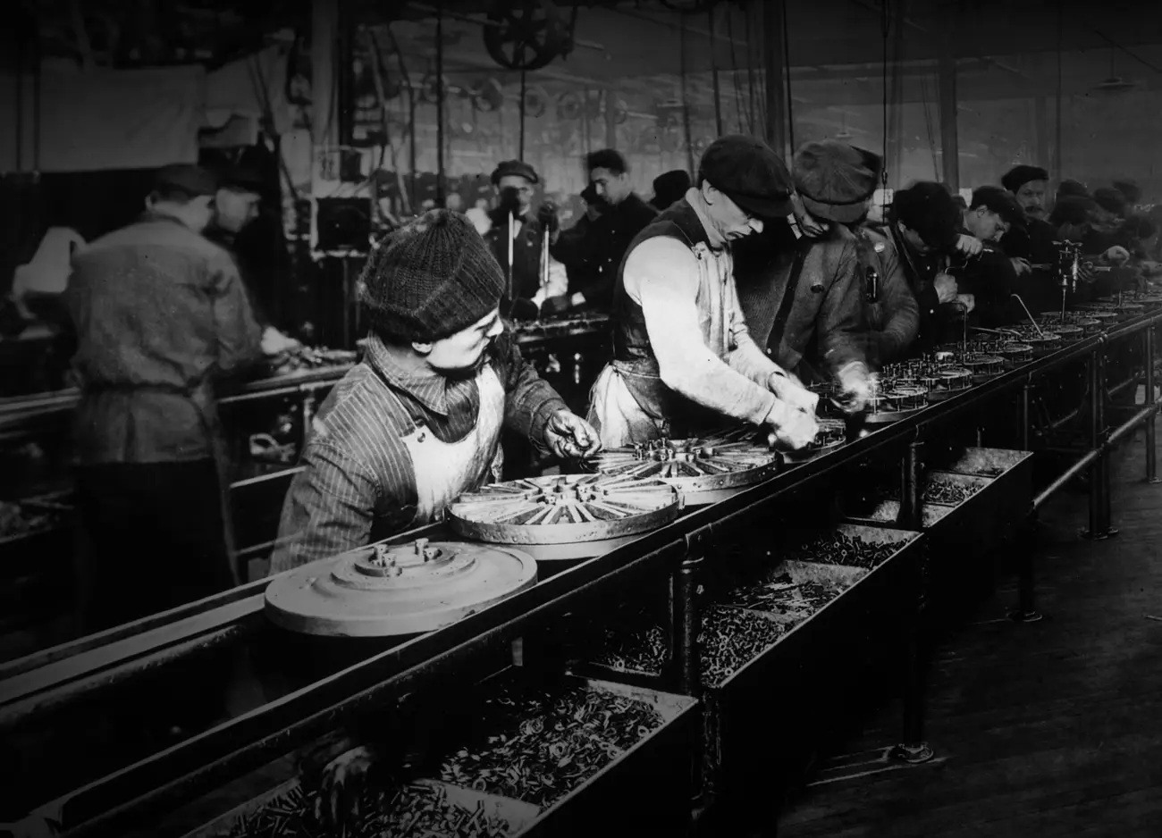 Une longue chaîne de montage dans un atelier de 1913 : des volants de magnéto posés sur un rail glissent d'un ouvrier au suivant ; chacun, penché, fait un seul geste, et des bacs de pièces s'alignent sous le rail.