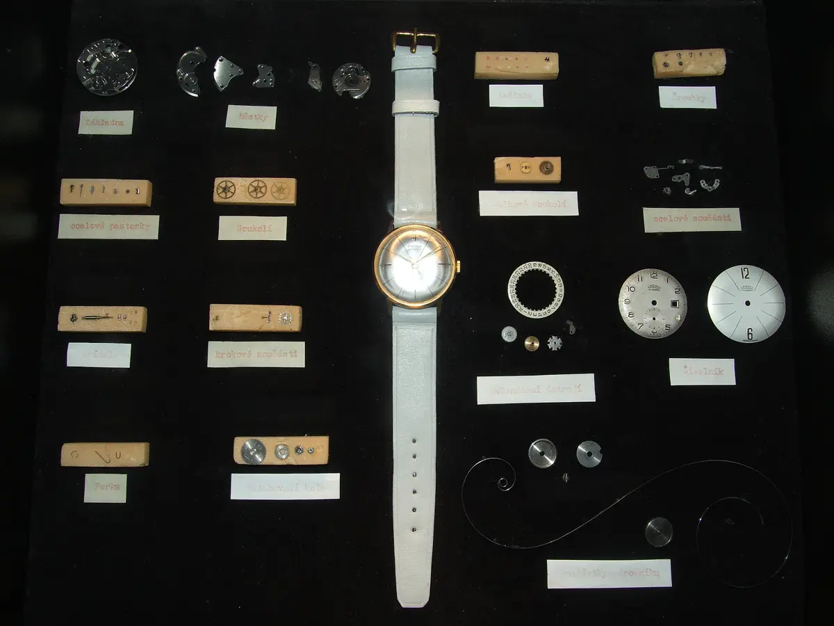 Une montre-bracelet posée au centre d'une vitrine, entourée de ses pièces détachées rangées par espèce sur de petits socles étiquetés : platine, ponts, pignons, roues, vis, ressorts, cadrans, spiral.