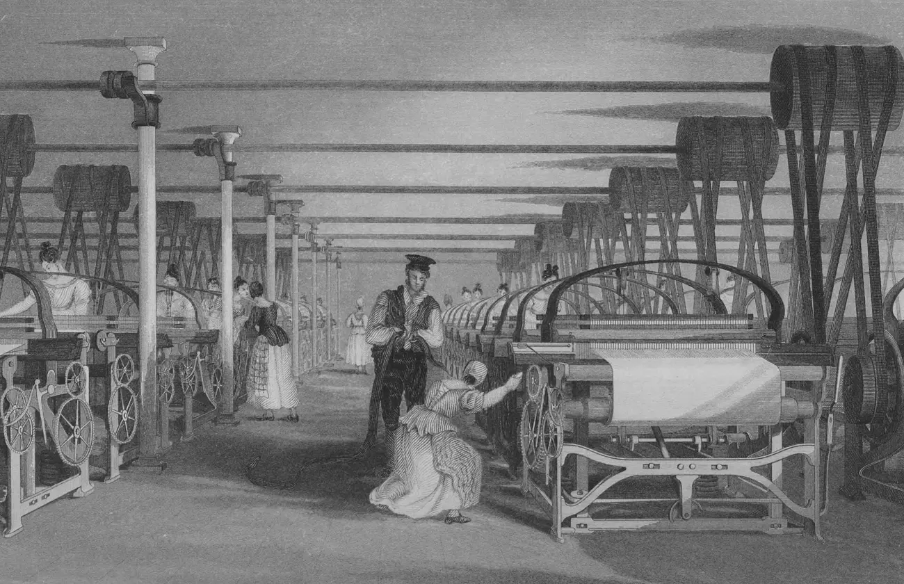 Une longue salle de métiers à tisser mécaniques, entraînés par des courroies qui descendent d'arbres de transmission au plafond ; des ouvrières surveillent les métiers, un contremaître est debout dans l'allée.